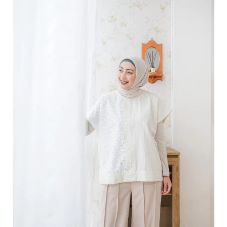 A woman wearing a cream-colored top by Abame with short sleeves and a round neckline, paired with trousers, standing in a room with a light-colored wall and a decorative mirror.
