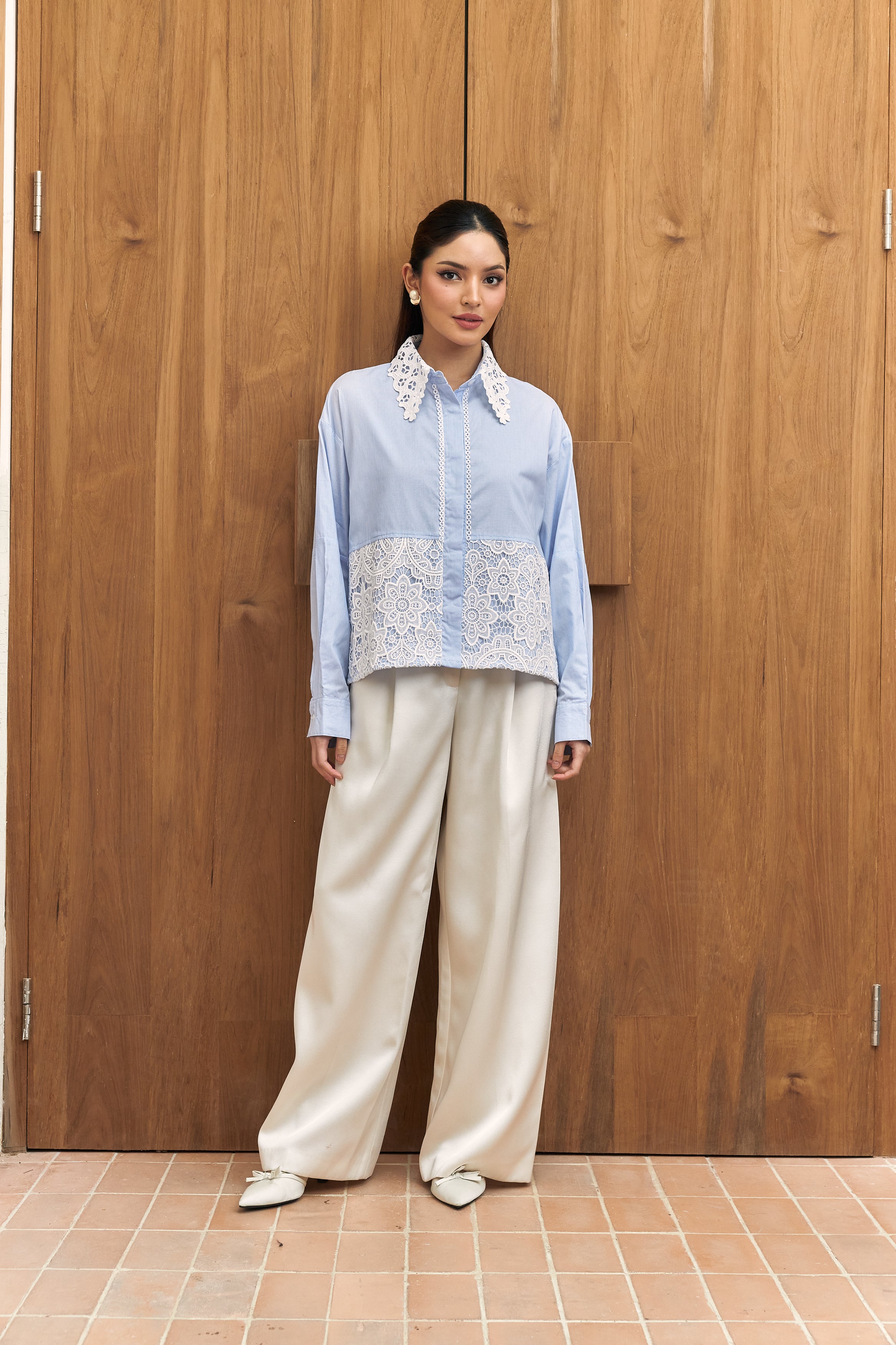 Woman wearing a light blue embroidered shirt by Studio Tui and beige pants standing against a wooden wall.