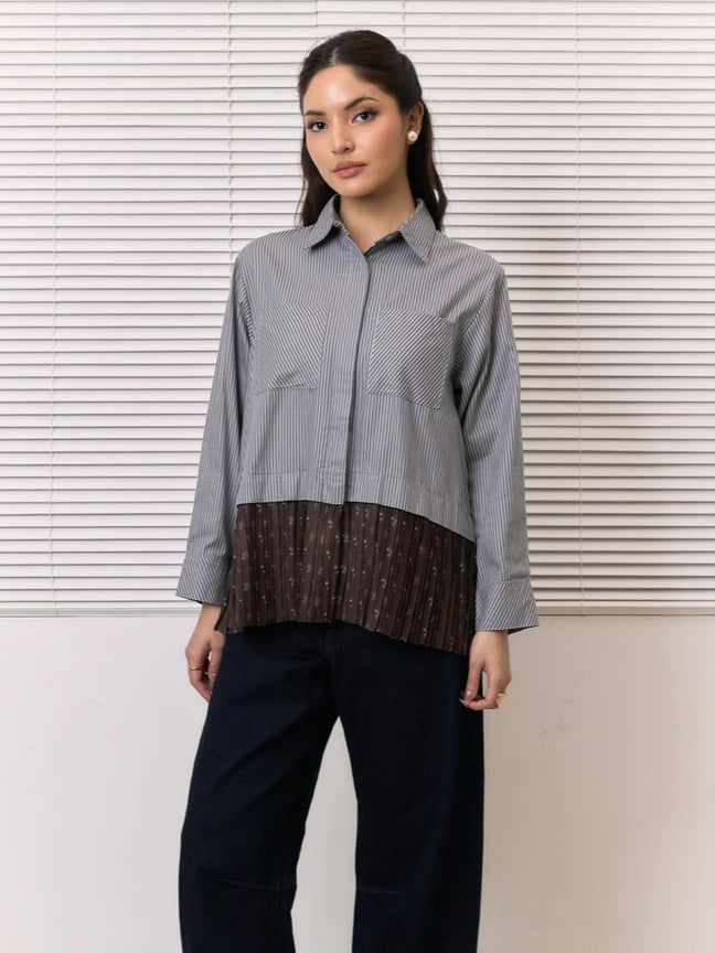 A woman modeling a long-sleeve button-up shirt by Studio Tui with a vertically striped gray and white upper section and a brown pleated lower section with small white floral motifs, paired with dark blue jeans, set against a background of white window blinds.