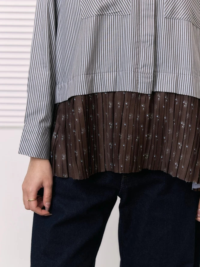 A close-up of a person wearing a long-sleeve shirt by Studio Tui with a vertically striped gray and white upper section and a brown pleated lower section with small white floral motifs, paired with dark blue jeans.