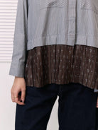 A close-up of a person wearing a long-sleeve shirt by Studio Tui with a vertically striped gray and white upper section and a brown pleated lower section with small white floral motifs, paired with dark blue jeans.