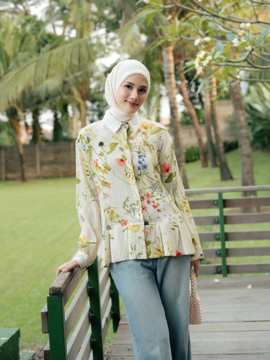 A woman wearing a cream floral long-sleeve blouse by Haidee Orlin with a white hijab and light blue jeans, standing on a wooden boardwalk in a park with green grass and palm trees in the background.