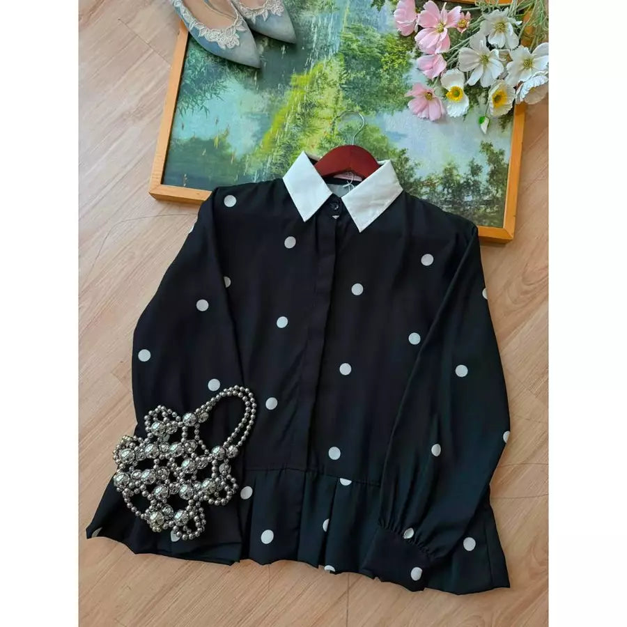 A black long-sleeve shirt by Haidee Orlin with white polka dots and a contrasting white pointed collar, laid flat on a light wood floor with decorative props including a pearl handbag, shoes, and a framed floral print.