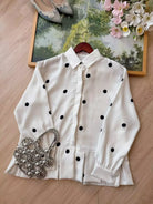 An off-white long-sleeve shirt by Haidee Orlin with black polka dots, styled on a wooden hanger on a light wood floor with a silver rhinestone purse, a pair of pointed shoes, and a floral picture frame in the background.