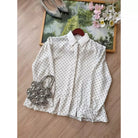 A white long-sleeve shirt by Haidee Orlin with a black polka dot pattern and a pointed collar, laid flat on a wooden floor with decorative accessories including a beaded handbag, shoes, and a framed picture.