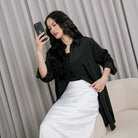 A woman wearing a black tunic over a black top and white pants, standing against a gray backdrop.