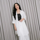 A woman wearing a white tunic over a black top and white pants, standing against a gray backdrop.