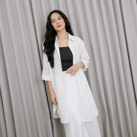 A woman wearing a white tunic over a black top and white pants, standing against a gray backdrop.
