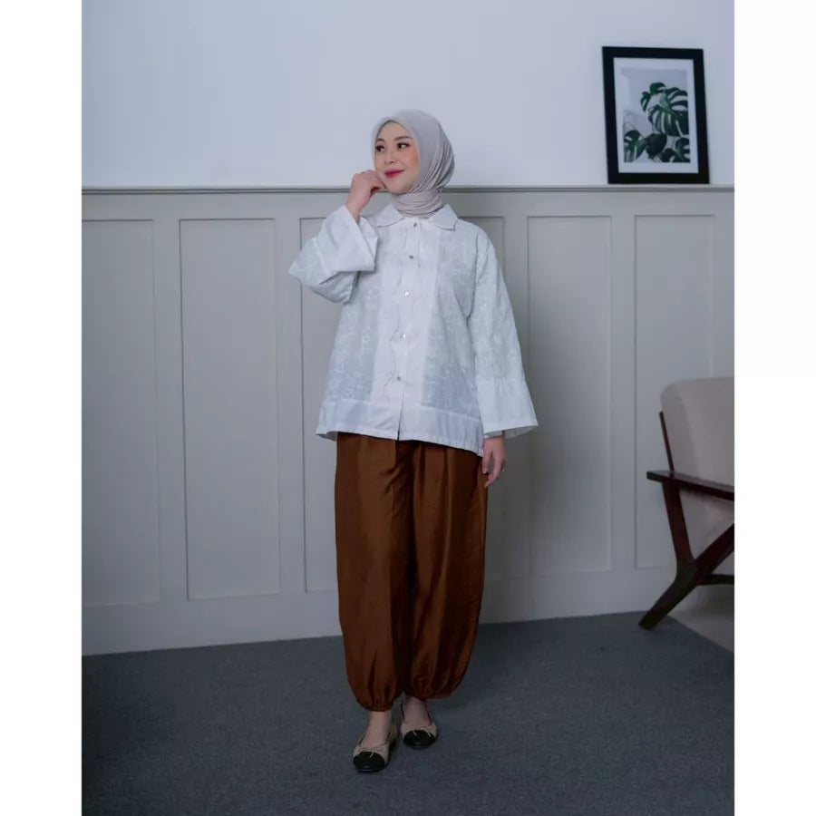 Woman wearing a white shirt by Abame and brown pants standing in a room with a white wall and a chair.