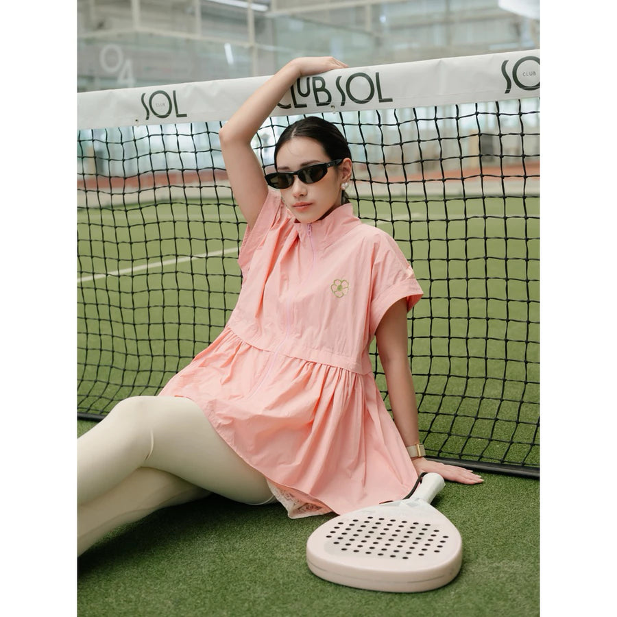 A woman wearing a light pink short-sleeve athletic vest by Haidee Orlin and beige leggings sits on a tennis court behind a net, holding a paddle. The dress has a small embroidered logo on the chest.