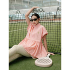 A woman wearing a light pink short-sleeve athletic vest by Haidee Orlin and beige leggings sits on a tennis court behind a net, holding a paddle. The dress has a small embroidered logo on the chest.