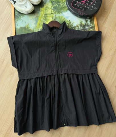 A black sleeveless zip-front long vest by Haidee Orlin with a gathered waist and a small pink paw-print logo on the chest, laid flat on a wood floor with shoes and a bag partially visible around it.