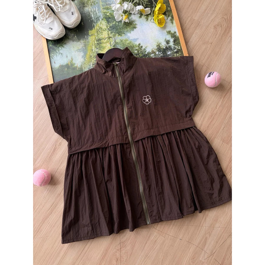 A dark brown sleeveless zip-front long vest by Haidee Orlin with a gathered waist seam, laid flat on a light wood floor. The garment has a small white embroidered logo on the chest and is styled with sneakers, flowers, and pink balls around it.