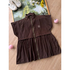 A dark brown sleeveless zip-front long vest by Haidee Orlin with a gathered waist seam, laid flat on a light wood floor. The garment has a small white embroidered logo on the chest and is styled with sneakers, flowers, and pink balls around it.