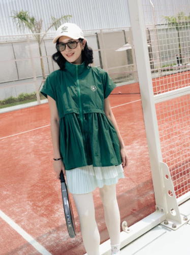 A woman stands on a red clay tennis court wearing a dark green short-sleeve zip-front vest by Haidee Orlin over a light green pleated mini skirt, with white leggings, a white cap, and sunglasses, holding a tennis racket.