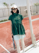 A woman stands on a red clay tennis court wearing a dark green short-sleeve zip-front vest by Haidee Orlin over a light green pleated mini skirt, with white leggings, a white cap, and sunglasses, holding a tennis racket.