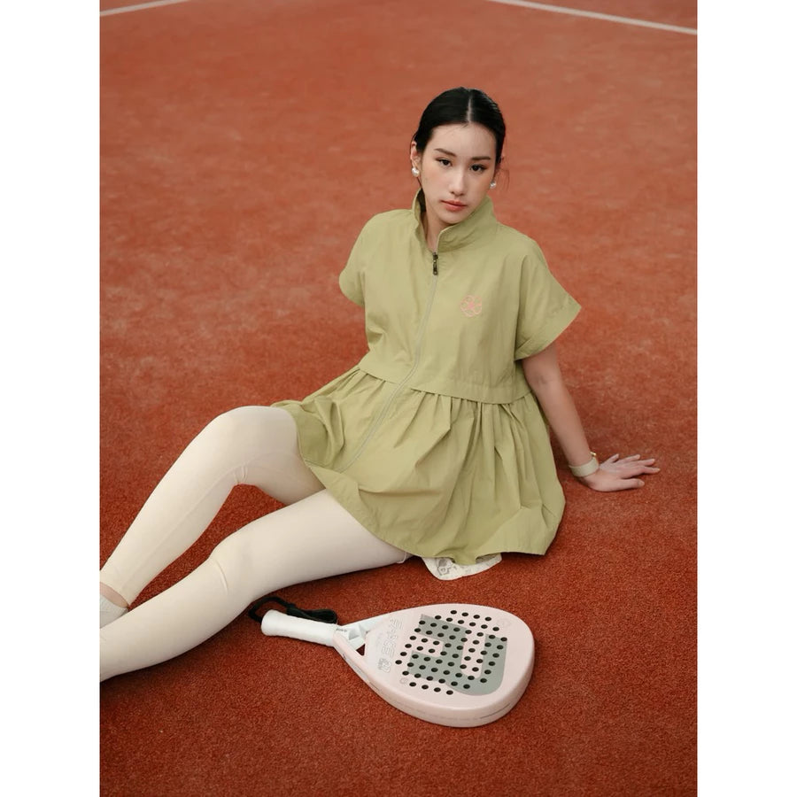 A woman sits on a clay tennis court wearing a light olive green short-sleeve athletic vest by Haidee Orlin and white leggings, with a paddle racket in front of her.