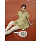 A woman sits on a clay tennis court wearing a light olive green short-sleeve athletic vest by Haidee Orlin and white leggings, with a paddle racket in front of her.