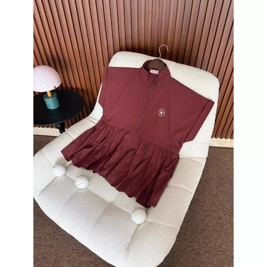 A burgundy short-sleeve zip-front longline vest by Haidee Orlin with a gathered ruffled hem, displayed on a cream boucle chair in an indoor setting with a slatted wood wall and a small table lamp.