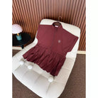 A burgundy short-sleeve zip-front longline vest by Haidee Orlin with a gathered ruffled hem, displayed on a cream boucle chair in an indoor setting with a slatted wood wall and a small table lamp.