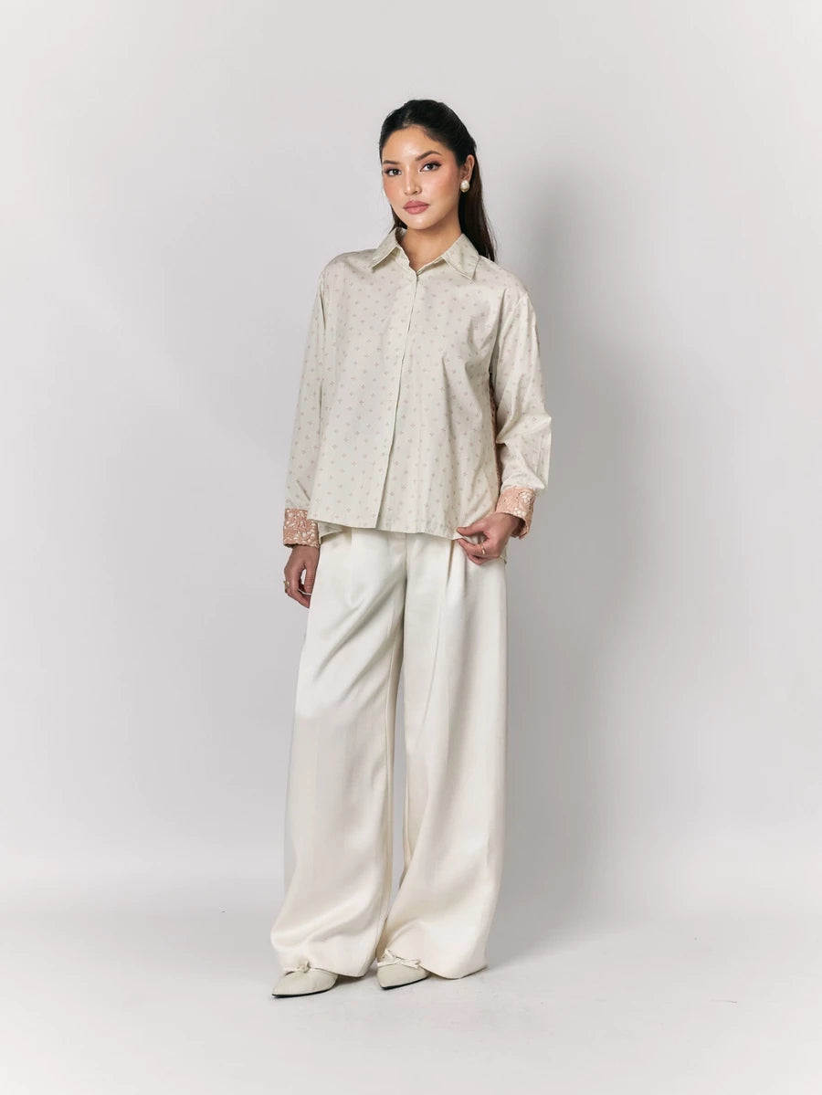 Woman wearing a beige shirt by Studio Tui and white pants against a plain background