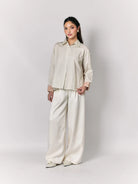 Woman wearing a beige shirt by Studio Tui and white pants against a plain background