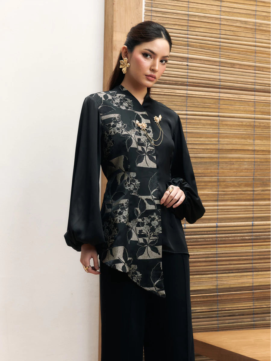 Woman wearing a black floral longsleeves blouse by Studio Tui against a wooden panel background