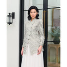 Woman wearing a sage green lace floral blouse by Kasa Heritage and white skirt standing in front of a glass door.