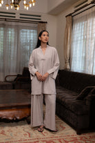 Woman in a light gray blouse by Kasyalia standing in a living room with curtains and furniture.