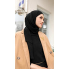 Woman wearing a black pashmina hijab by Puru Kambera and beige coat with a brand logo, standing outdoors.