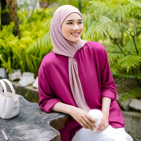 Woman in a pink outfit with a pashmina hijab by Puru Kambera sitting outdoors holding a white cup.
