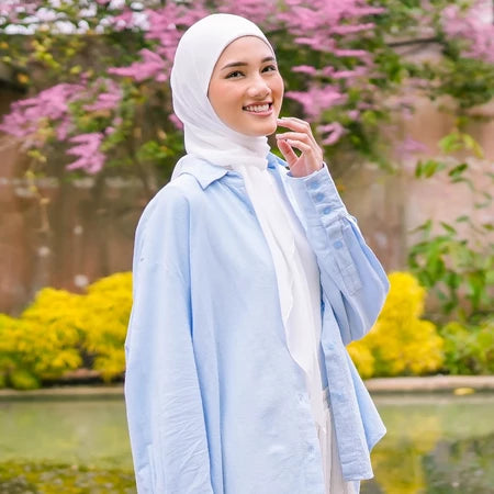 Woman wearing a light blue jacket and white pashmina hijab by Puru Kambera standing in front of pink flowers and greenery.