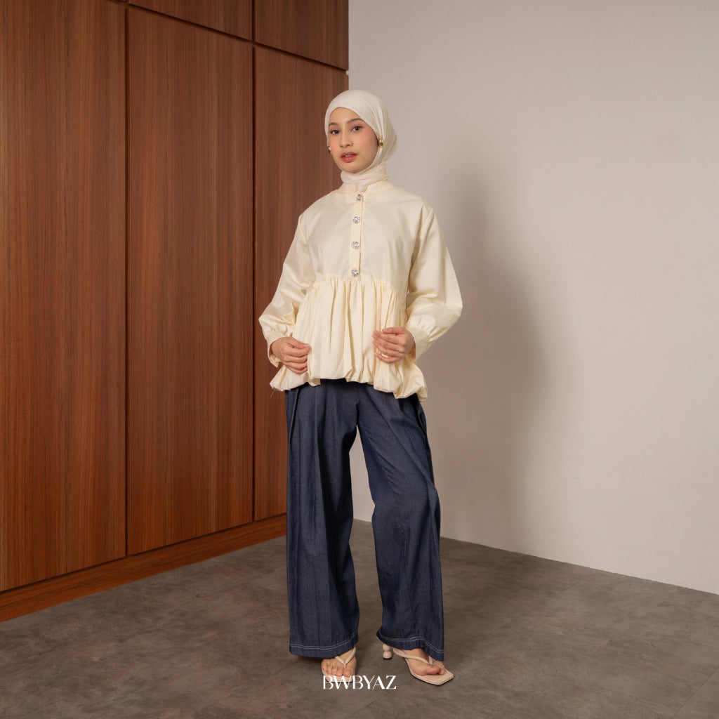 Woman wearing a cream puff blouse from BWBYAZ and navy pants against a wooden panel background