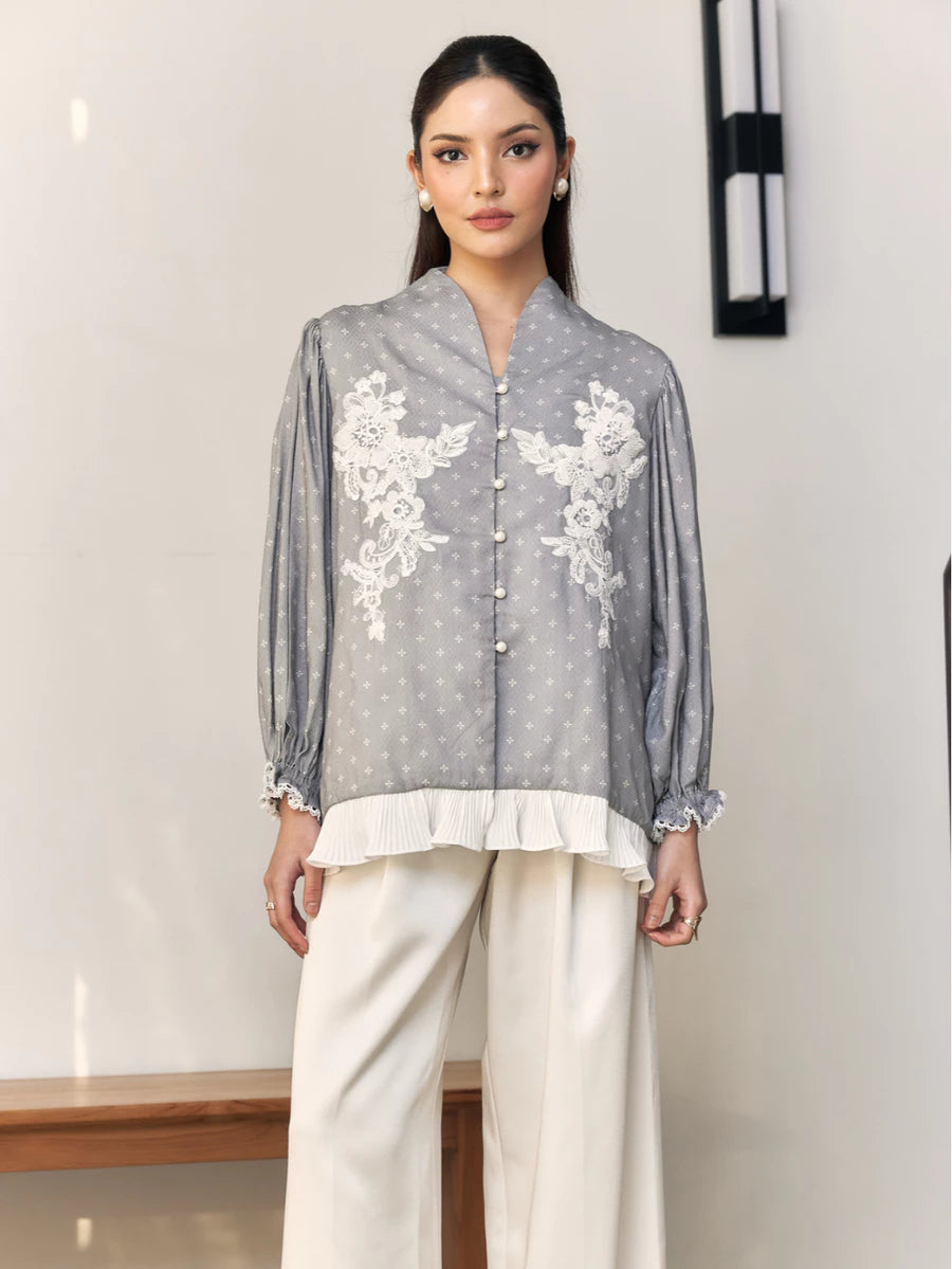 Woman wearing a gray blouse with white floral embroidery by Studio Tui and white pants indoors.