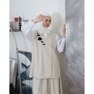Woman wearing a white hijab, white shirt, and beige pinstripe vest by Maima and pants in an indoor setting.