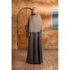 Person wearing a beige hijab and gray Mecca Dhurma Abaya by Nehhat Modest standing in front of a wooden door.