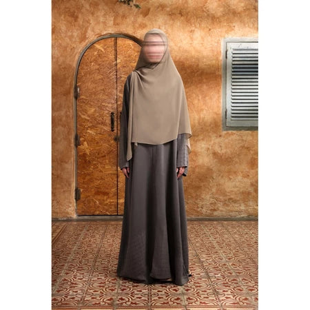 Person wearing a beige hijab and gray Mecca Dhurma Abaya by Nehhat Modest standing in front of a wooden door.