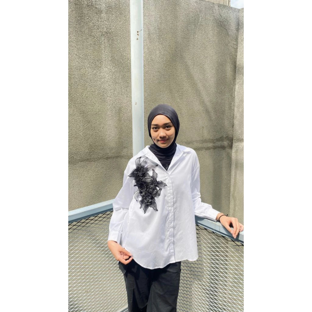 Person wearing a white shirt with black floral design from Maima and dark pants standing against a textured wall.