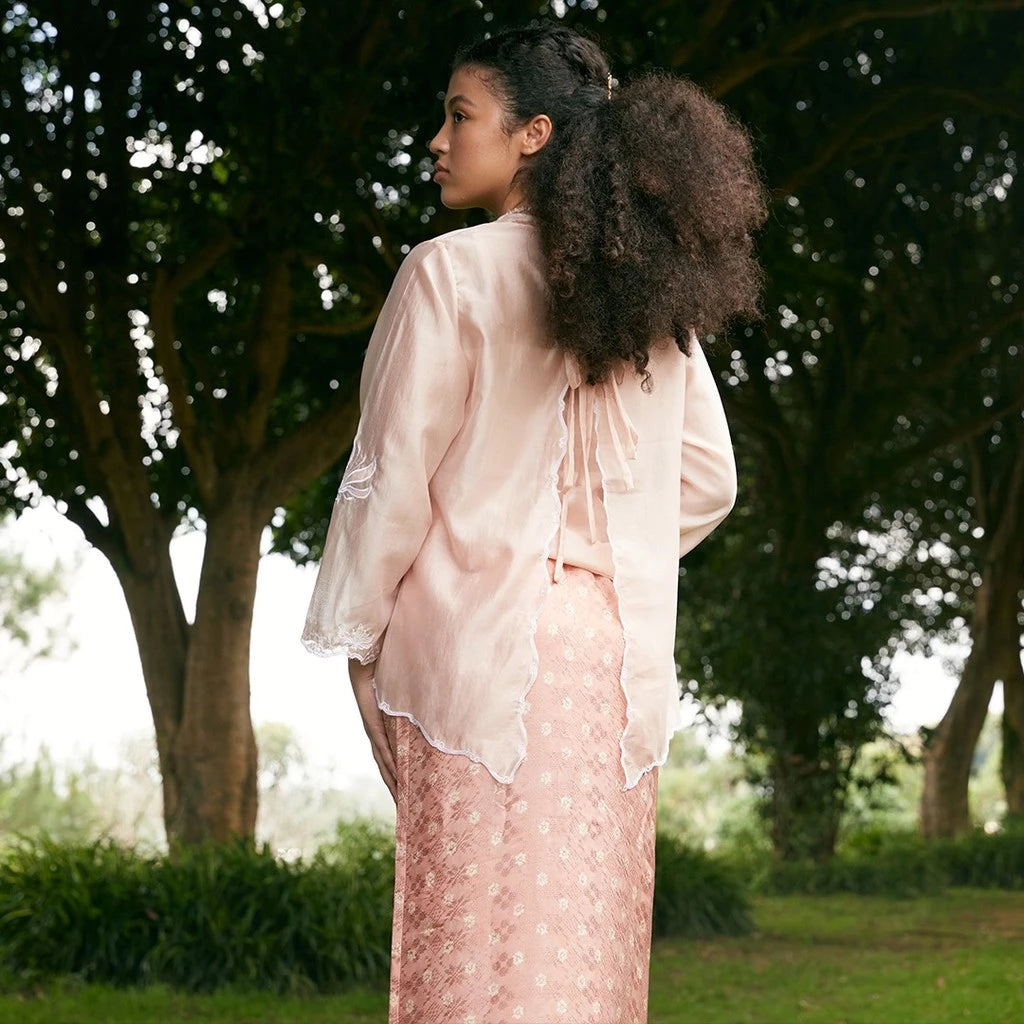Woman in a peach colored outer by Kartinis Label standing in a park with trees in the background