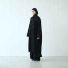 Person wearing a long black coat by Rashawl and headscarf on a white background