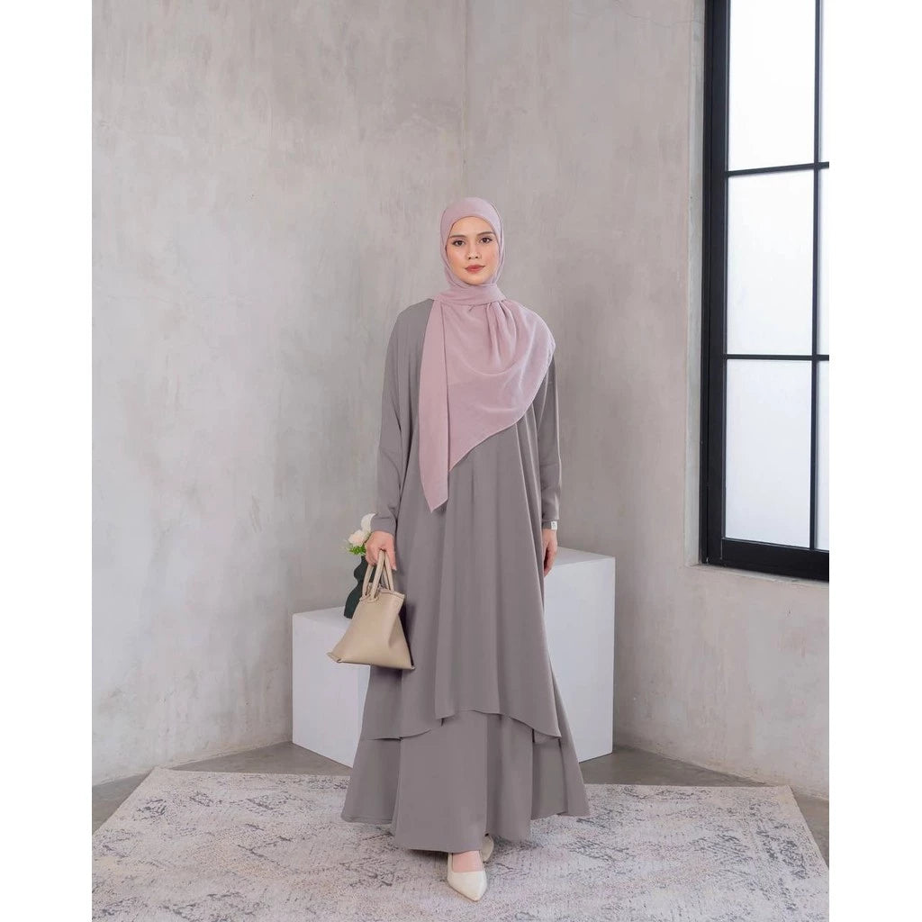 A woman wearing a dark brown tunic dress with a hijab, standing in a room with minimalist decor.