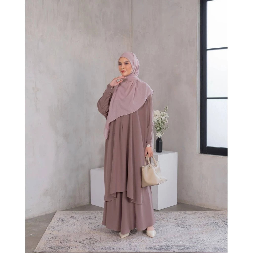 A woman wearing a purple tunic dress with a hijab, standing in a room with minimalist decor.