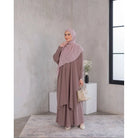 A woman wearing a purple tunic dress with a hijab, standing in a room with minimalist decor.