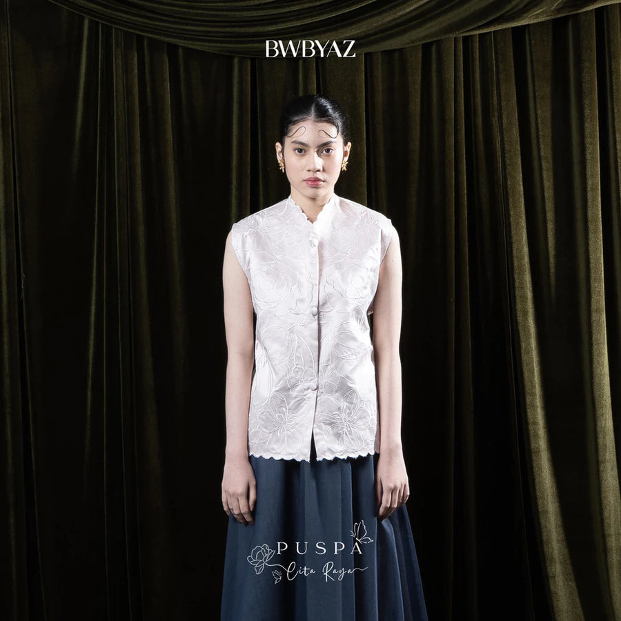 Woman wearing a light pink sleeveless embroidered vest and dark skirt against a dark curtain background with 'BWBYAZ' branding.