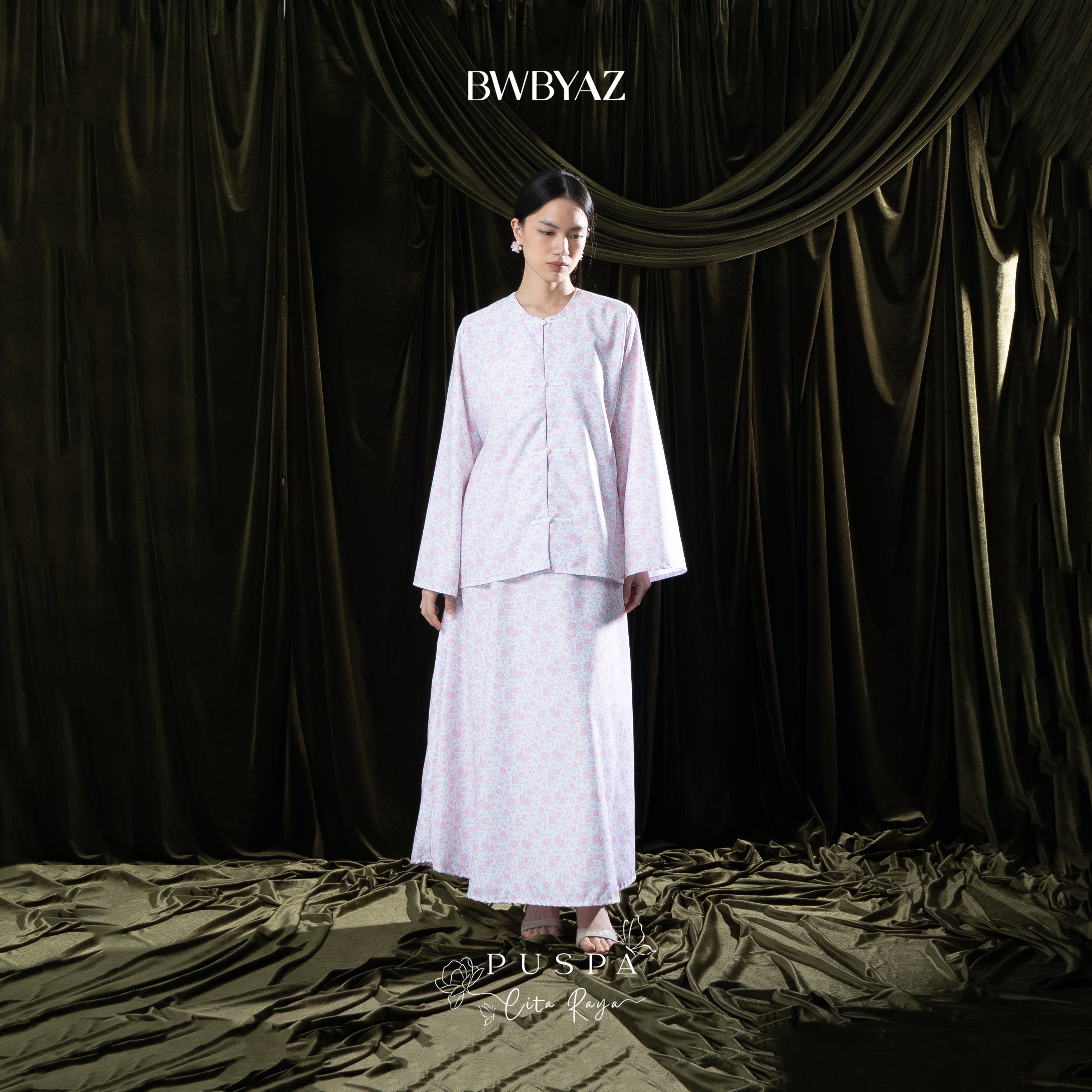 Woman wearing a light pink traditional outfit with 'BWBYAZ' branding against a dark curtain background.