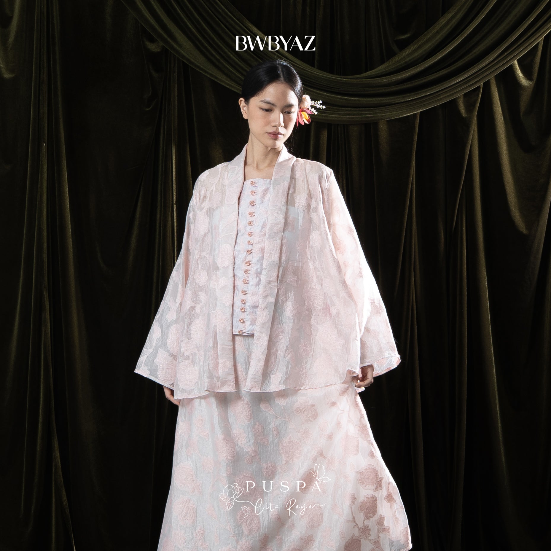 Woman wearing a light pink kebaya with lace details on a dark curtain background, featuring the brand 'BWBYAZ'.