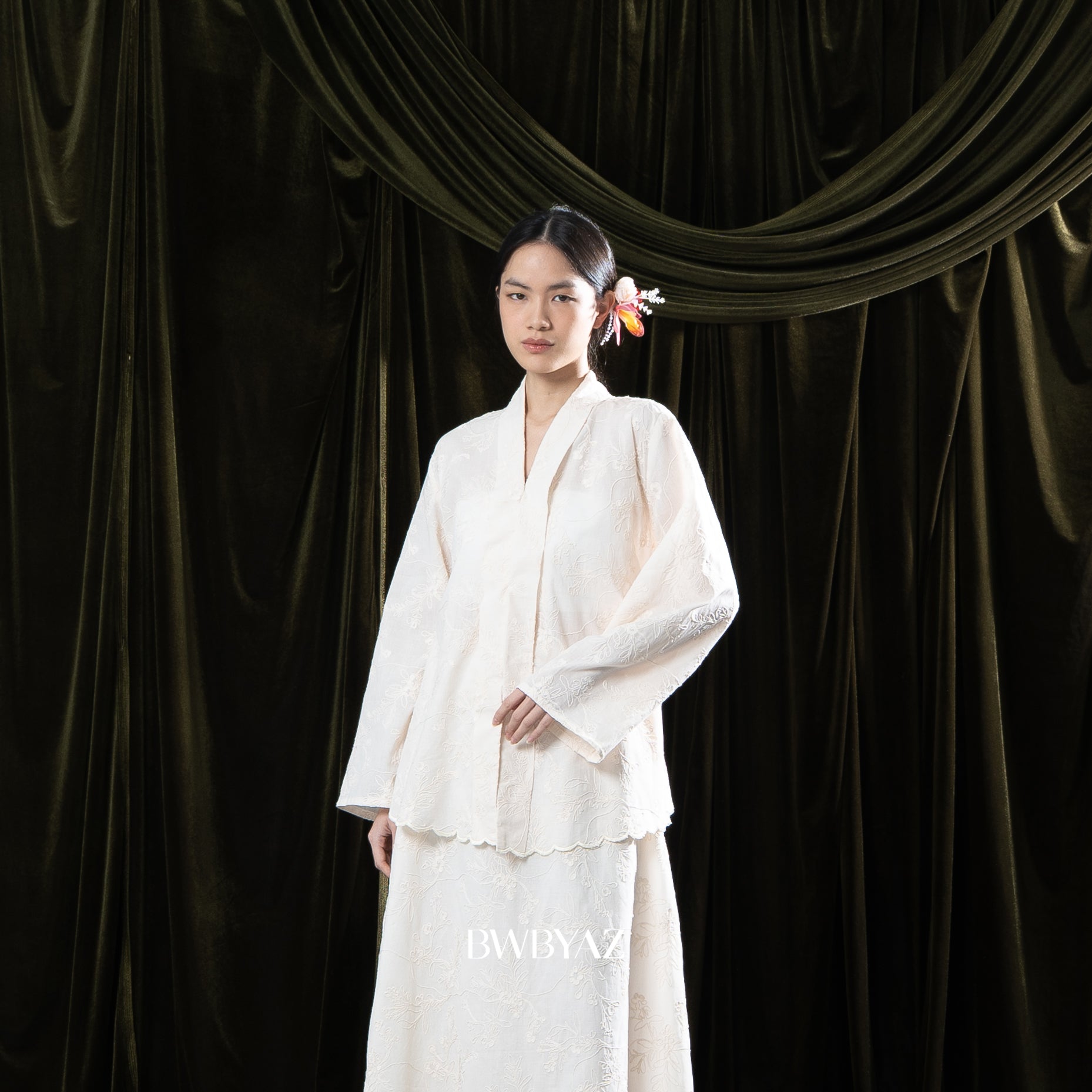Woman in a cream traditional dress standing against a dark green curtain with 'BWBYAZ' branding.

Seorang wanita mengenakan kebaya berwarna krem berdiri di depan tirai hijau gelap dengan merek ‘BWBYAZ.