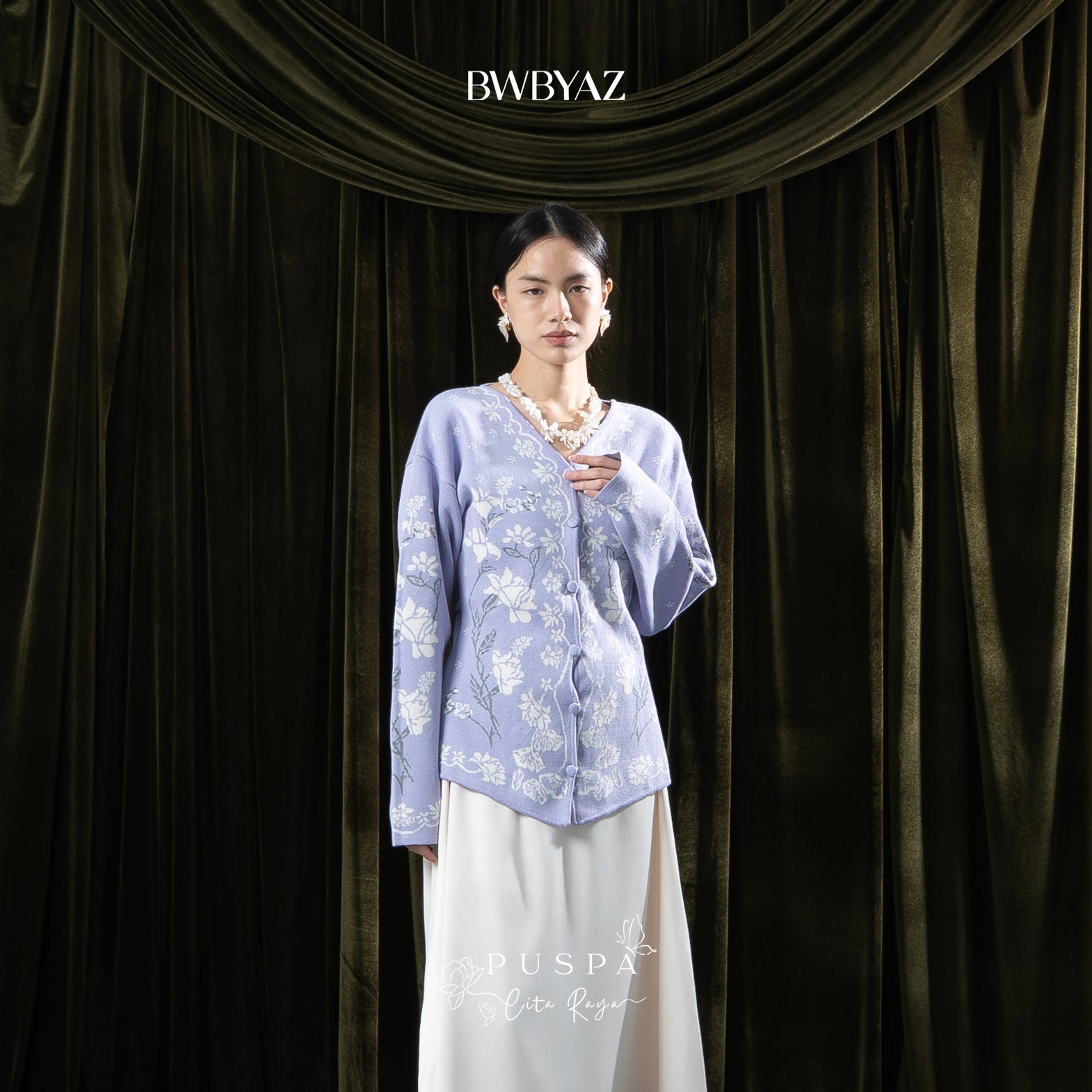Woman wearing a lavender-colored floral cardigan and white skirt against a dark curtain backdrop with 'BWBYAZ' branding.
