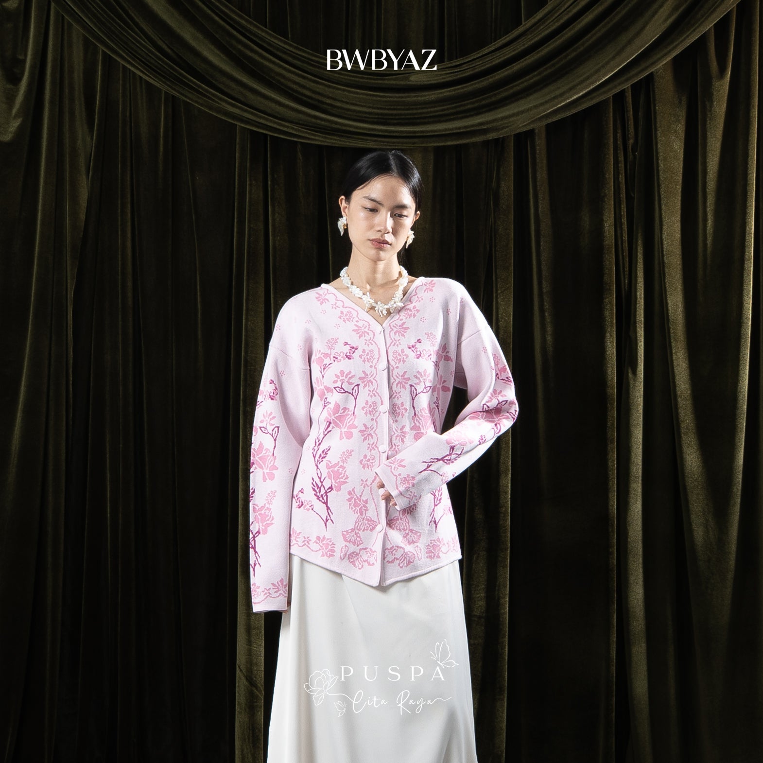 Woman wearing a pink floral cardigan and white skirt against a dark curtain backdrop with 'BWBYAZ' branding.