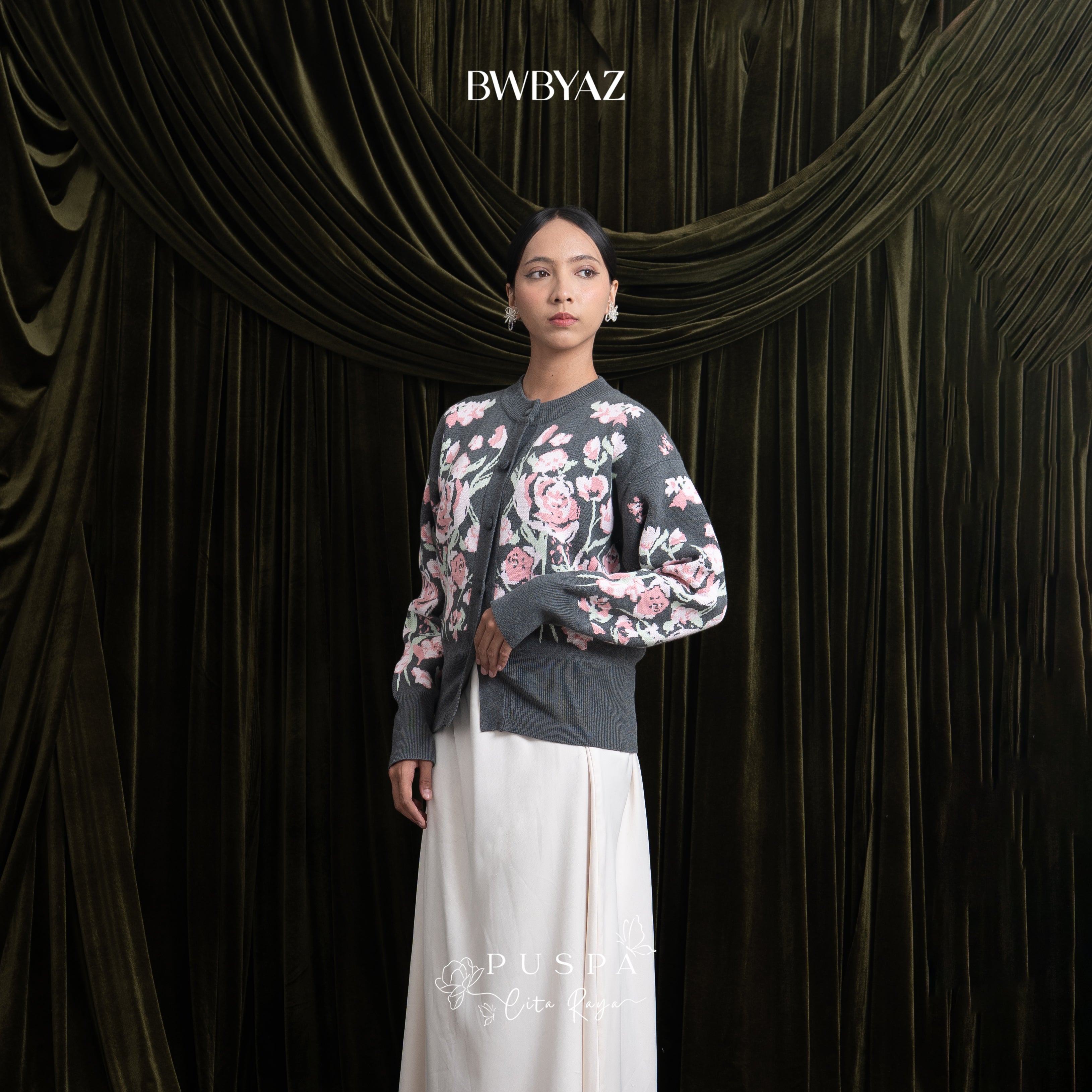 Woman wearing a grey floral cardigan and white skirt against a dark curtain backdrop with 'BWBYAZ' branding.